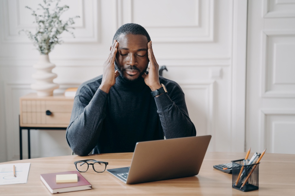 Concerned young african american man employee suffering from headache at workplace, sitting with closed eyes and frustrated face expression, worried male office worker thinking of problem solution. Concerned young african american man employee suffering from headache at workplace