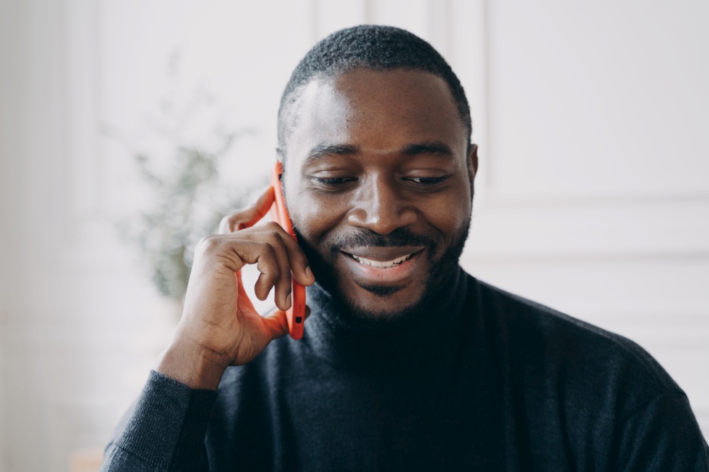 Close up of young smiling african man having business conversation on mobile phone while working in office, cheerful dark-skinned guy holding smartphone talking with friend or consulting client. Young smiling african man having business conversation on mobile phone