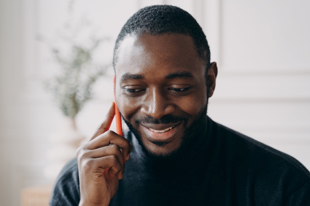 Cheerful young African American businessman talking on smartphone while working distantly in modern home office interior. Mobile communication and freelance, male entrepreneurship concept. Cheerful young African American businessman talking on smartphone while working distantly at home
