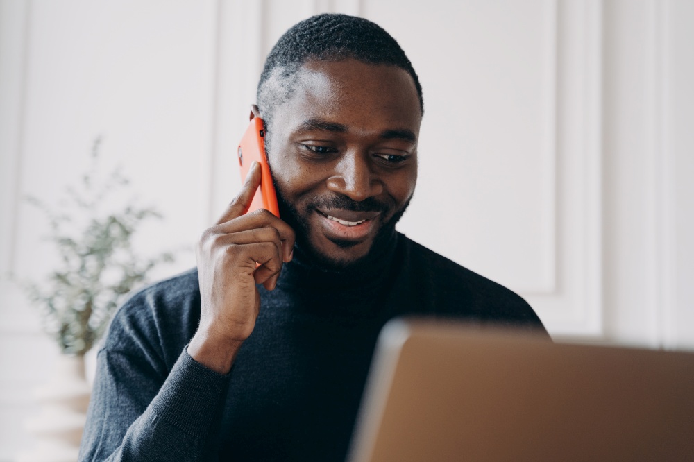 Cheerful young man with African lineage Ecommerce business owner calling to supplier for confirmation of orders while working remotely online from home office comparing and checking products on laptop. Cheerful young man with African lineage commerce businessowner calling to supplier for confirmation