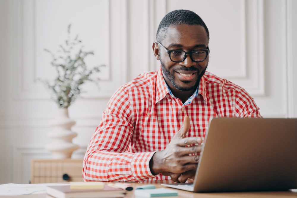 Young smiling african american businessman in glasses having pleasant conversation with business partner by video call on laptop, discussing work results during online meeting with colleagues. Young smiling african american businessman talking with business partner by video call on laptop