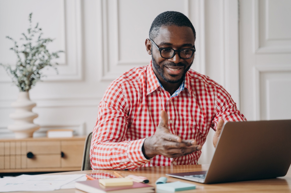 Positive african man online teacher tutor looking at laptop screen and smiling, talking with students while working remotely from home, dressed in casual clothes. E-learning and distant education. Positive african man online teacher tutor looking at laptop screen and talking with students