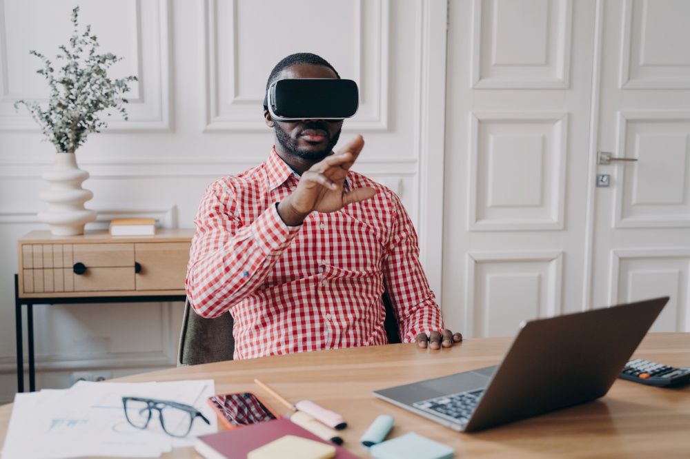 Young African ethnicity man entrepreneur in VR headset glasses immersed in 3D gaming in virtual reality relaxing during remote working online sitting at desk at modern home office in front of laptop. Young African ethnicity man entrepreneur in VR headset glasses immersed in 3D gaming