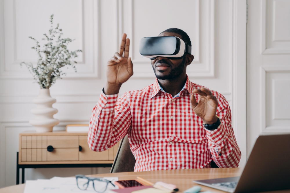 Focused Aframerican man in VR glasses enjoying augmented reality while sitting at workplace at home office, pointing with fingers up in air interacting with virtual 3D objects, using electronic device. Focused Aframerican man in VR glasses enjoying augmented reality while sitting at workplace