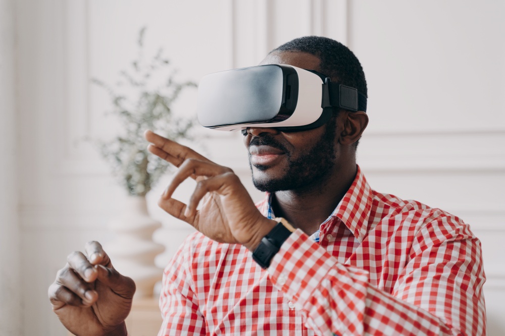 Young african businessman wearing VR glasses touching 3d objects in air with hands while playing games or watching video in digital world, interacting with interface in virtual reality at work. Young african businessman wearing VR glasses touching 3d objects, interacting with virtual reality