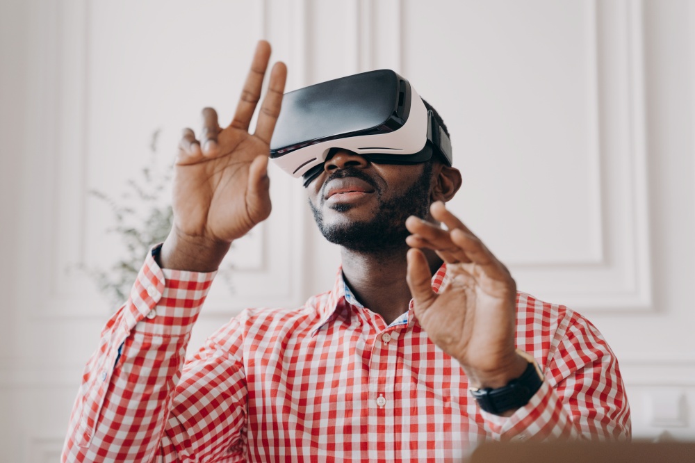 Black man in VR glasses experiencing 3D technology of augmented reality. Excited African male waving with hands up and down makes touching moves with fingers, stands against home office background. Black man in VR glasses experiencing 3D technology of augmented reality