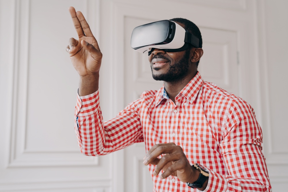Enthusiastic smiling african ethnicity guy in VR headset glasses enjoying virtual reality online trying cyber world simulation at work, raised hands touching air with fingers, having fun with 3D gadget. Impressed african ethnicity guy in VR headset glasses enjoying virtual reality online at work