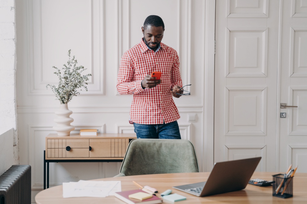 Young serious african american businessman in casual clothes standing in office with smartphone, using mobile phone, reading online news or chatting with client on cellphone, making call at work. Young serious african american businessman in casual clothes standing in office with smartphone
