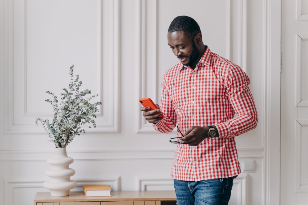 Attractive Afro American businessman in casual wear holding smartphone trying to answer important call. Confident mixed race man entrepreneur works remotely from modern home office. Freelance concept. Attractive Aframerican businessman in casual wear holding smartphone trying to answer important call