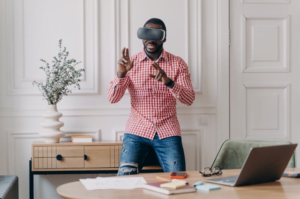 Young programmer or software developer African American man in VR headset glasses testing 3D games and applications standing in modern home office interior. Technology and software development concept. Young programmer or software developer African American man in VR headset glasses testing 3D games