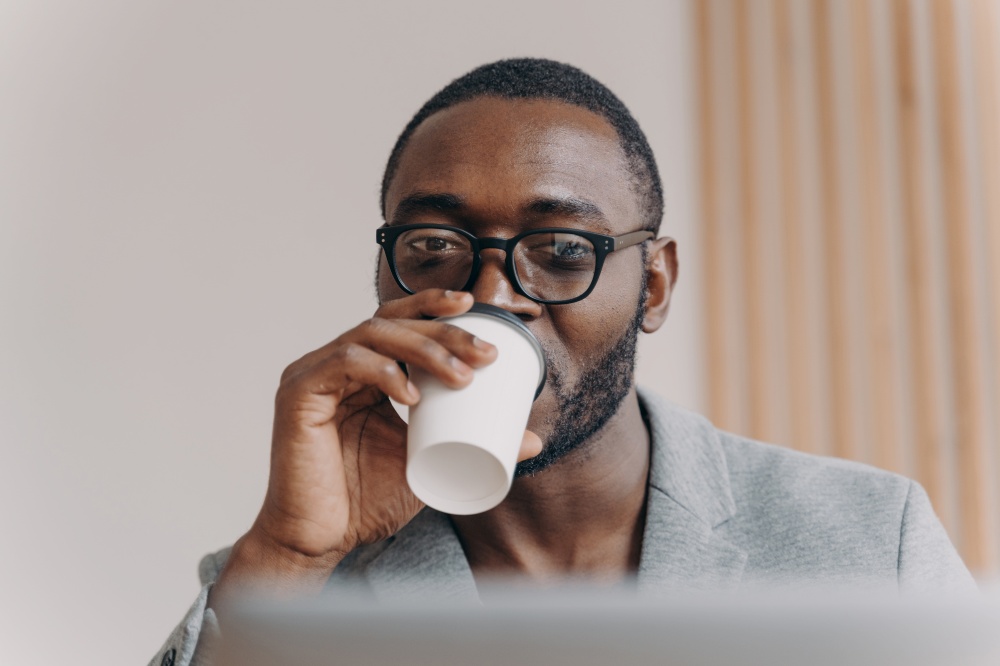 Attractive confident Afro american man company CEO drinking take away coffee from disposable cup while looking at laptop screen following remote financial meeting online during video conference. Attractive confident Aframerican man company CEO drinking coffee while looking at laptop screen
