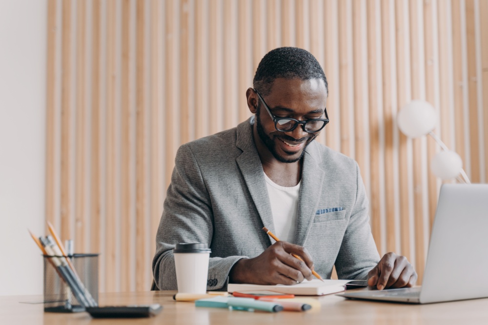 Positive elegant african male employee having video conference online with colleagues, smiling american businessman makes notes in agenda while sits at desk at home office and using laptop. Happy afro american smiling businessman makes notes in agenda while having video call online