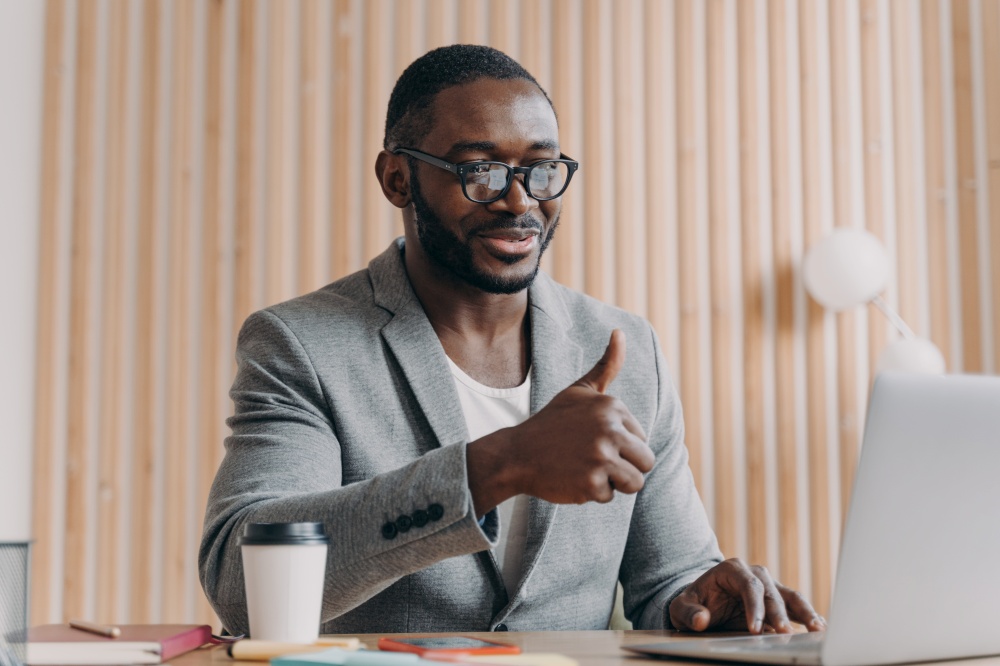 Happy young african american male employee in glasses talks online on laptop with client or business partner, showing thumb up and smiling, cheerful dark-skinned businessman making video call at work. Happy young african american businessman talks online on laptop with client or business partner