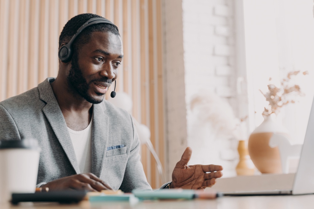 Young smiling african american man in headset consulting client customer via video call on laptop, pleasant dark-skinned guy foreign language tutor giving online lesson talking with students. Young smiling african american man in headset consulting client customer via video call on laptop