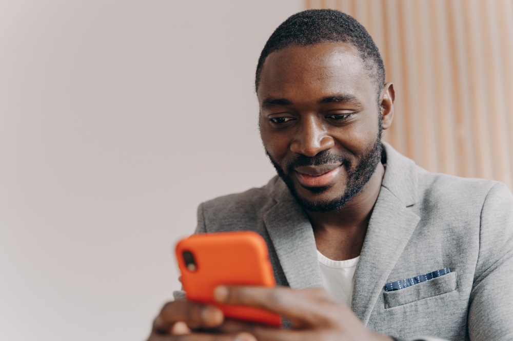 Attractive smiling young African self-employed man in formal suit chatting with partners on smartphone, satisfied businessman sending emails to company office before oncoming business meeting. Attractive confident young Afro american self-employee chatting with partners on smartphone
