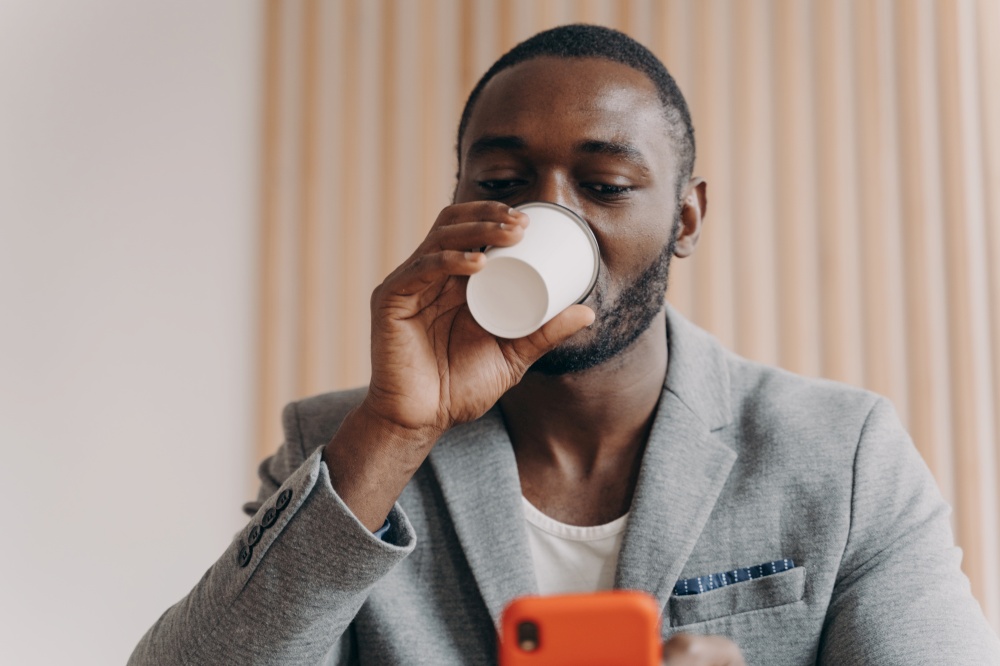 Elegant young African ethnicity businessman in modern blazer drinking take away coffee from disposable cup while looking on cellphone screen checking important and urgent emails from company office. Elegant African ethnicity businessman drinking take away coffee while looking on cellphone screen