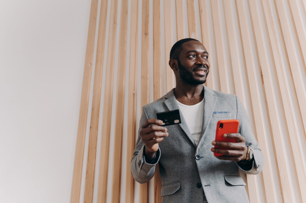 Excited African-American businessman in stylish blazer using smartphone and credit card for shopping online, satisfied black man ordering food online, paying for long-awaited purchase via e-banking. Excited African-American businessman using smartphone and credit card for shopping online