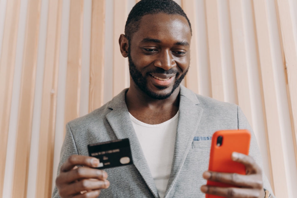 Happy african businessman in suit holding credit card and mobile phone while buying goods or services via Internet, smiling afro american man using payment e-banking system at work. Happy african businessman in suit holding credit card and mobile phone while doing online shopping