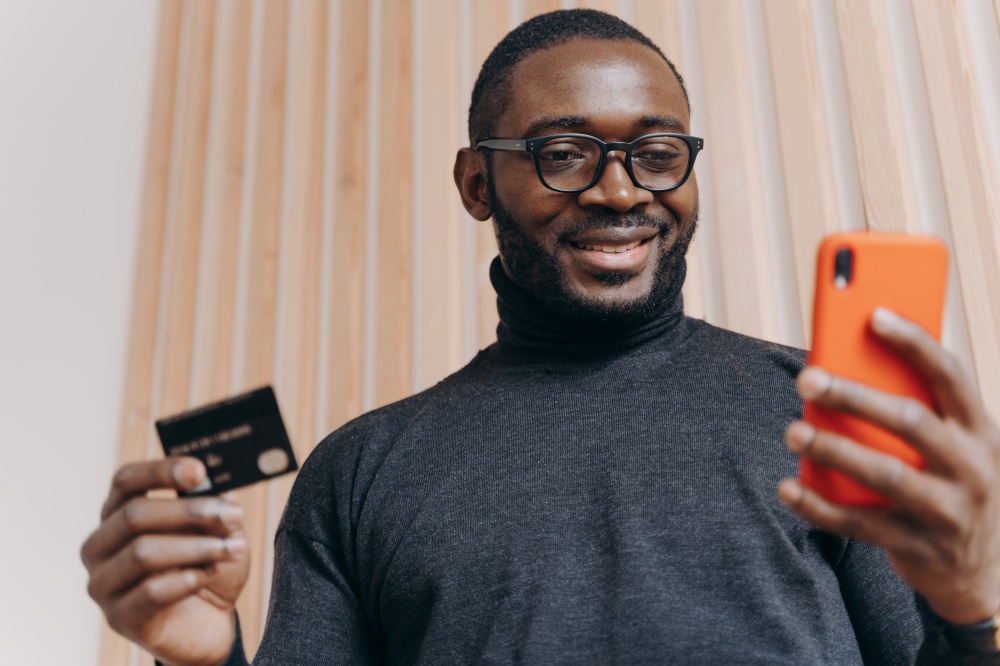 Online payment. Cheerful African american businessman holding credit card and smartphone for shopping in internet, with happy smile looking on phone screen satisfied with successful transaction. Cheerful African american businessman holding credit card and smartphone for shopping in internet