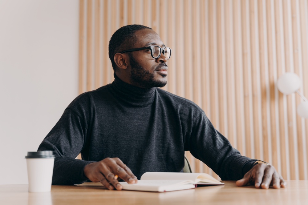 Young thoughtful african american businessman sitting at desk with pen and agenda looking away with pensive expression. Millennial biracial entrepreneur coming up with new ideas at home office. Thoughtful african businessman sits at desk with pen and agenda looks away with pensive expression