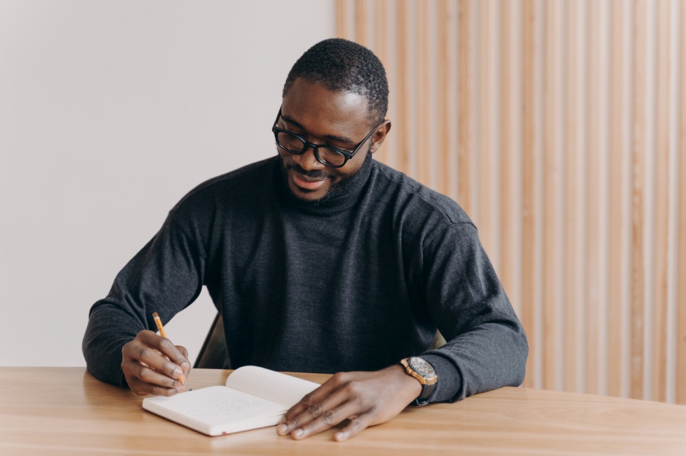 Young african american man entrepreneur in glasses taking notes in agenda while sitting at desk at home or office,planning workday, writing down some fresh ideas or work schedule. Young african american man entrepreneur in glasses taking notes in agenda