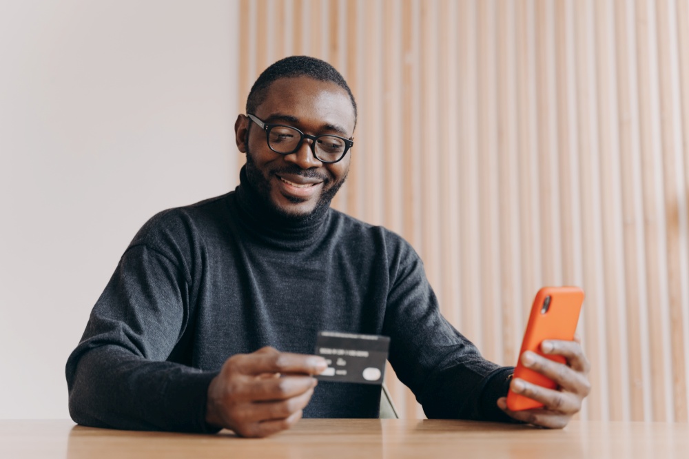 Cheerful Aframerican man entrepreneur holding credit card and using touchscreen smartphone for online shopping while making orders online sitting at home office. Ecommerce and online payment concept. Cheerful Aframerican man entrepreneur holding credit card and smartphone for online shopping