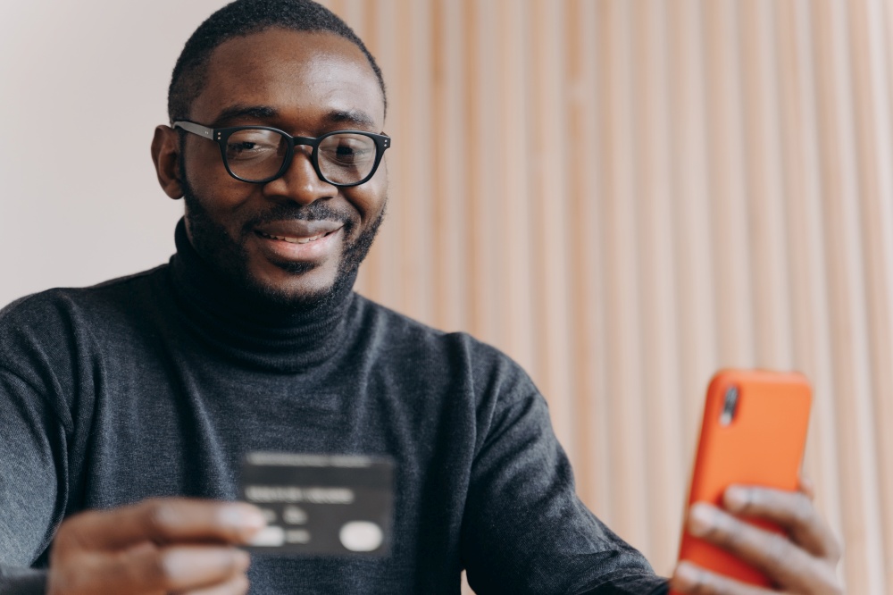 Positive African ethnicity man entrepreneur in glasses paying with credit card online while making order on cellphone. Smiling millennial guy making transaction using mobile bank application. Young positive African ethnicity man entrepreneur in glasses paying with credit card online