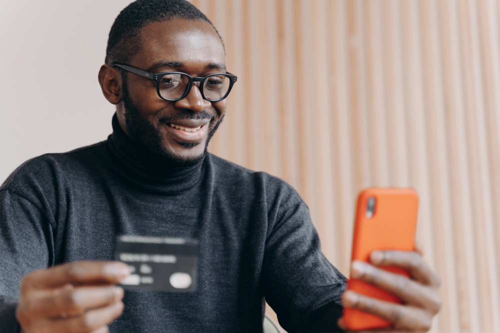 Smiling young African American businessman using credit card and smartphone while sitting at workplace in modern office.Cheerful employee doing online purchase or contacts with banking support service. Smiling African American businessman using credit card and smartphone while sitting at workplace