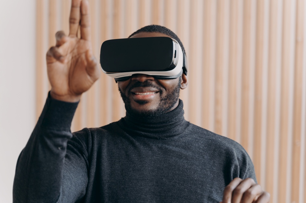 Excited African American man in VR headset glasses enjoying augmented reality while sitting at modern workplace at home, cheerfully raised hand with two fingers up as interacting with cyberspace in 3D. Excited African American man in VR headset glasses enjoying augmented reality in office