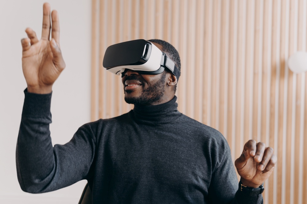 Young smiling african american businessman in VR glasses sitting in office, testing virtual reality goggles for business, impressed man using 3D technology, touching objects in air with hands. Smiling african american businessman in VR glasses sits in office, testing virtual reality goggles