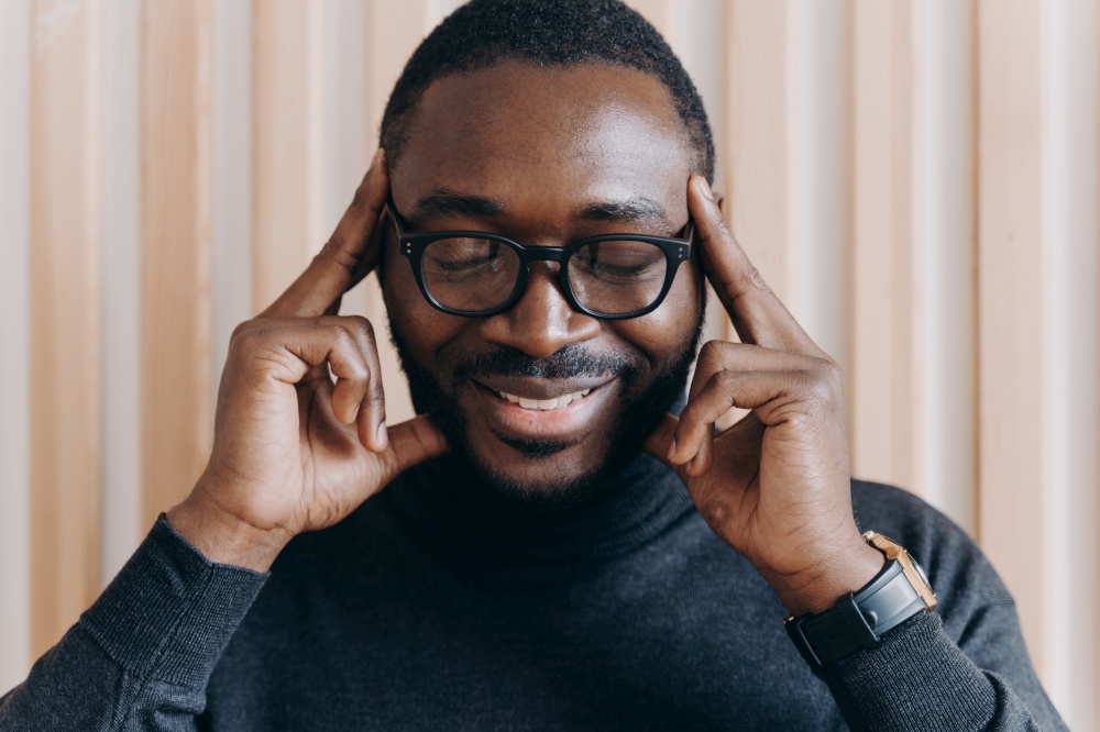 Smiling thoughtful African American male entrepreneur posing with closed eyes and with forefingers touching temples, showing brooding expression and birth of new ideas and successful projects. Thoughtful African American male entrepreneur with closed eyes touching temples with forefingers