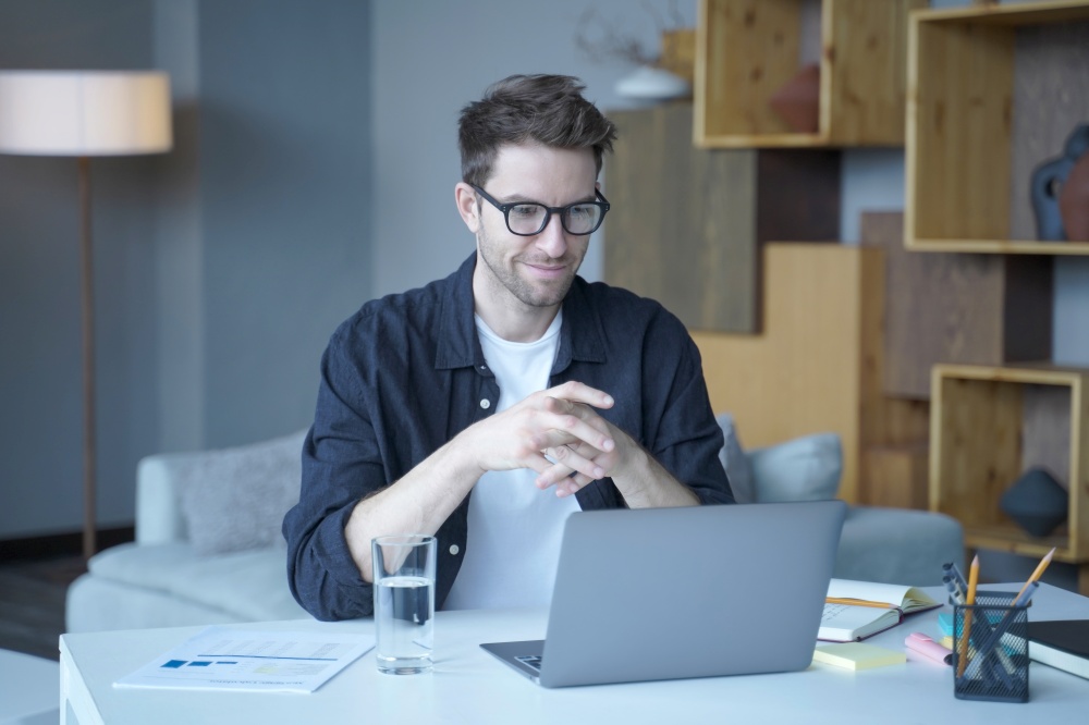 Young handsome german man wearing glasses working on laptop computer remotely from home, smiling male freelancer in casual clothes sitting at workplace during remote work. Freelance concept. Young handsome german man wearing glasses working on laptop computer remotely from home