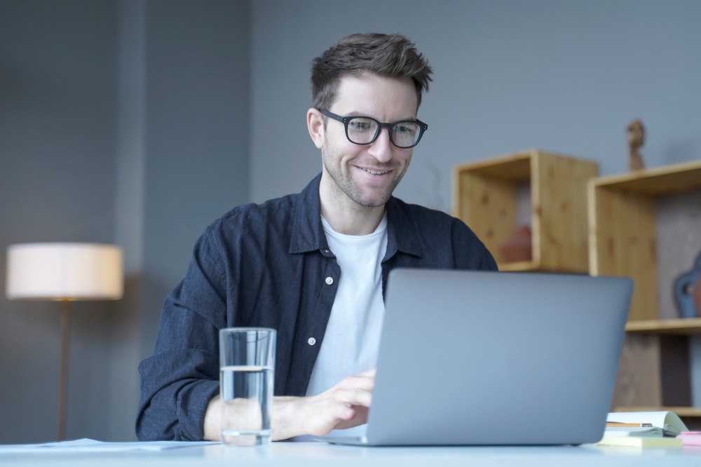 Young smiling businessman European financial consultant working remotely from home, chatting online with colleagues about details from last meeting, typing on laptop keyboard while looking at screen. Young smiling businessman European financial consultant working remotely online from home office