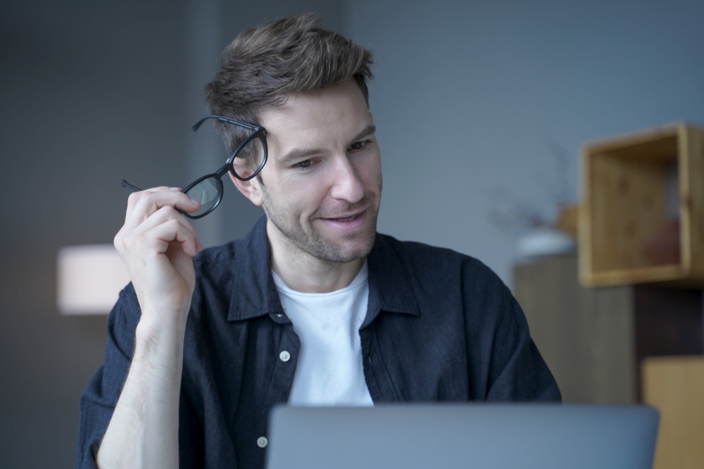 Attractive positive businessman in casual wear took off his glasses, remotely working from home office on new company project, focused European entrepreneur looking at laptop screen with head tilted. Attractive positive businessman in casual wear took off his glasses, remotely working from home
