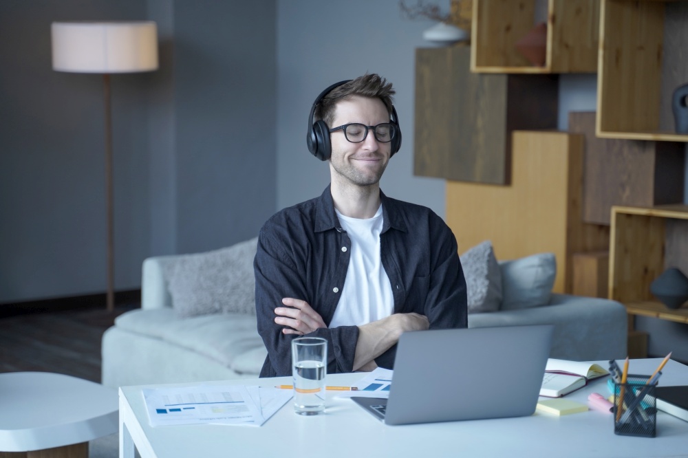 Young smiling happy austrian male freelancer sitting with closed eyes in headphones and enjoying listening to music at home office, man listens podcasts or audiobooks during remote work on laptop. Young happy austrian male freelancer in headphones enjoying listening to music at home office