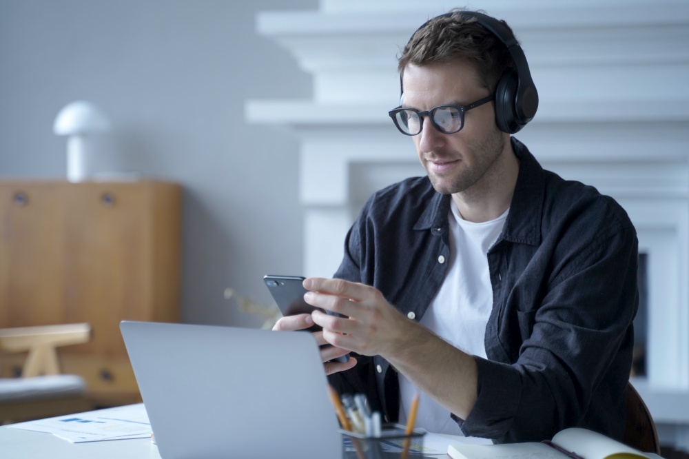 Serious austrian guy freelancer in headphones using phone at workplace, sitting at desk at home office working on laptop and at same time responding on important email or sms on smartphone in hands. Pensive male entrepreneur in headphones sitting at desk at home office and using mobile phone