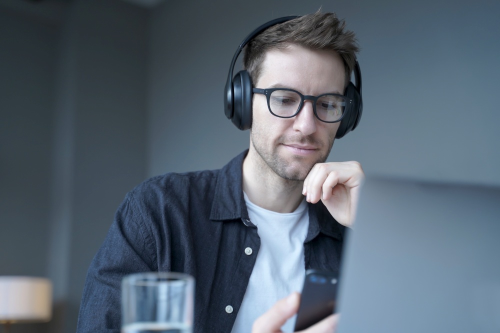 Young focused german man in glasses wearing wireless headphones using mobile phone, listening music at workplace in office, watching funny internet videos on smartphone during work break. Young focused german man in wireless headphones using mobile phone listening music at workplace