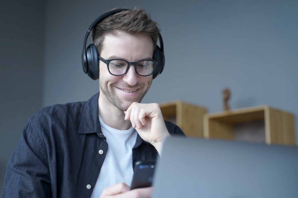 Attractive happy German home office worker in headphones and glasses sincerely smiling when looking at mobile phone screen in his hands while working remotely on laptop online. Freelance concept. Happy home office worker in headphones and glasses sincerely smiling when looking at phone screen