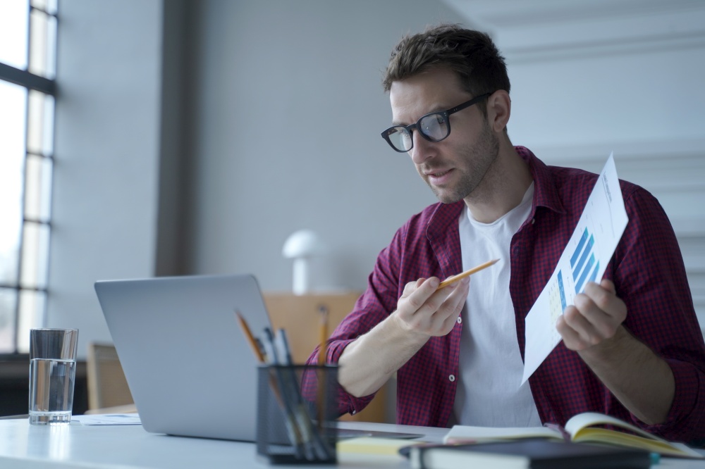Young european man office worker in glasses showing statistics report during online meeting with business partner, german guy financial advisor consulting client remotely from home, using laptop. Young european man office worker in glasses showing statistics report during online meeting