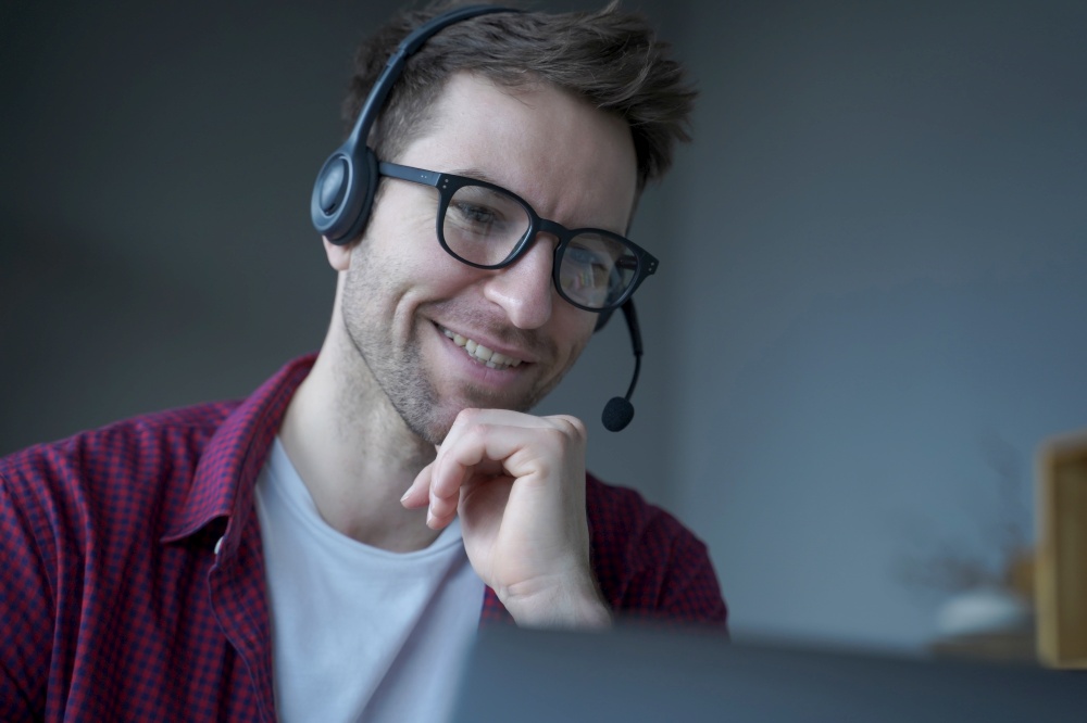 Cheerful man German speaking callcenter consultant in headset with mic works remotely from home, politely smiling while communicates online via laptop with customers, helps in solving current problems. Cheerful man German speaking call center consultant in headset with mic works remotely from home