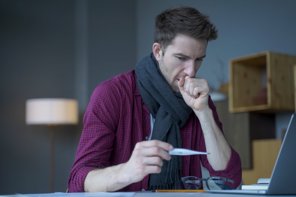 Unhealthy looking young male entrepreneur in warm knitted scarf around his neck coughing looks at digital thermometer in his hand not believing that caught flu while working remotely online from home. Unhealthy looking male entrepreneur in scarf around his neck coughing looks at digital thermometer