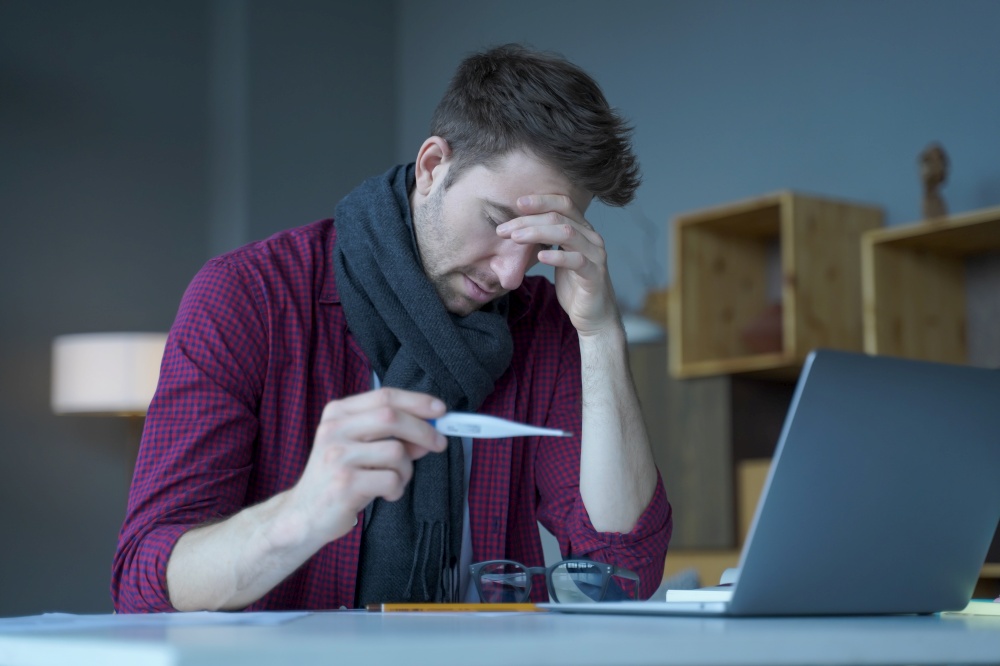 Unhappy austrian man freelancer dressed in warm scarf checking body temperature at work, holding thermometer with worried face expression, suffering of covid or flu symptoms at workplace. Unhappy austrian man freelancer checking body temperature at work, holding thermometer