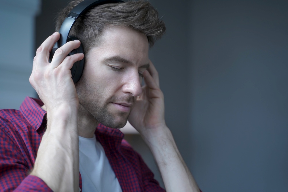 Side view photo of young pleased german man with closed eyes listening music in headphones with delighted face expression, austrian guy enjoying favourite playlist while spending free time at home. Young pleased german man with closed eyes listening music in headphones at home with delighted face