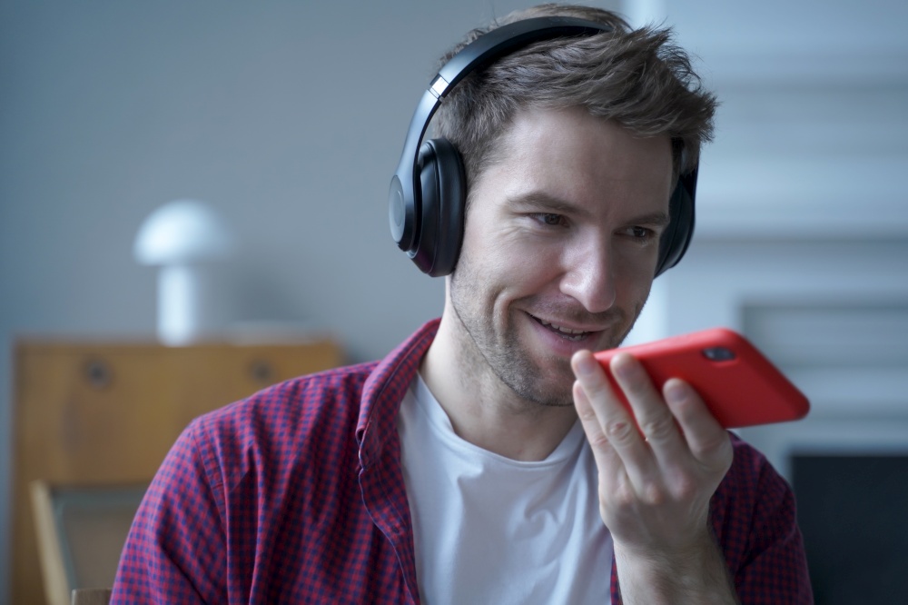Young smiling german guy in headphones using translator app while studying online, holding mobile phone and talking on speakerphone with friend, recording voice message while spending time at home. Young smiling german guy in headphones using translator app on mobile phone while studying online