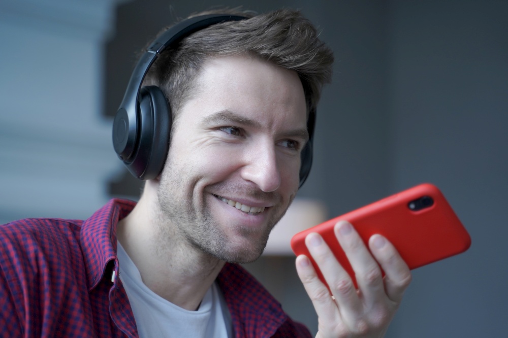 Close up of cheerful young german man in wireless headphones holding smartphone in hand with microphone close to his face during mobile conversation with friend, recording audio message. Cheerful young german man in wireless headphones holding smartphone in hand, recording audio message