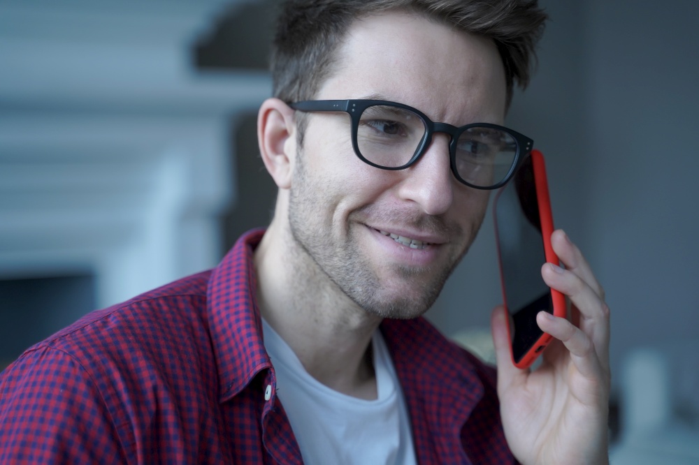 Young smiling german guy in glasses using mobile phone and talking on with friend while spending leisure free time at home, happy man holding smartphone and making call to client during remote work. Young smiling german guy in glasses using mobile phone and talking on with friend