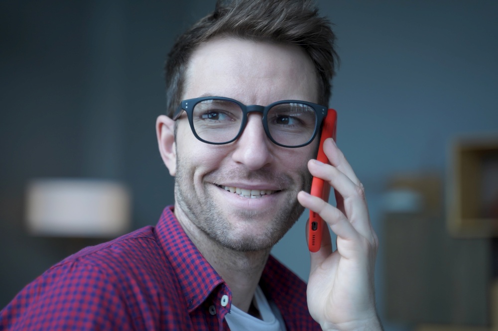 Close up photo of young handsome cheerful german man in glasses making phone call, talking on smartphone with client or business partner, pleasant austrian businessman enjoying mobile conversation. Close up photo of young handsome cheerful german man in glasses making phone call