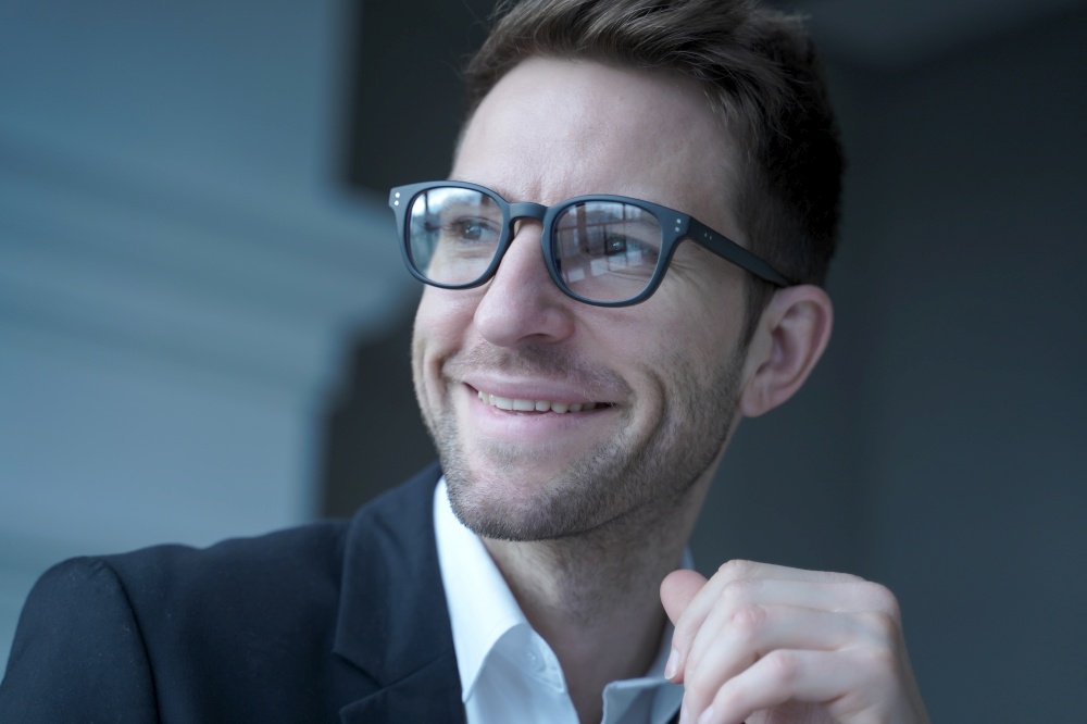 Close up portrait of positive young male entrepreneur in glasses and formal wear looking aside with pleasant smile distracted from remote work on computer in office. Freelance and business concept. Positive young male entrepreneur in glasses and formal wear looking aside with pleasant smile