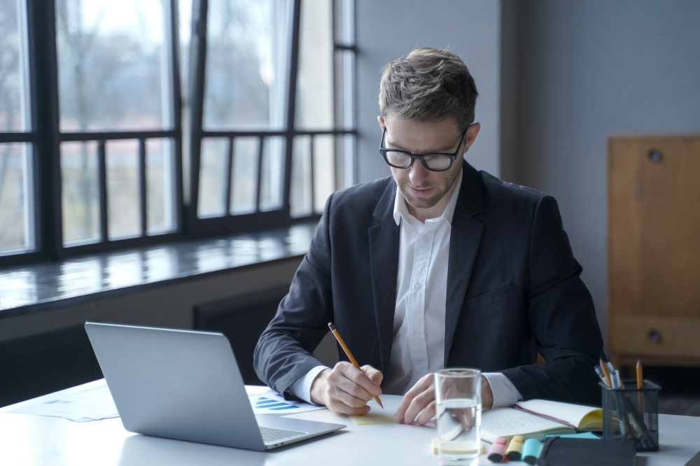 Immersed in work. Focused young German male banker writes down memos on sticky notes to remember important details to share for upcoming online meeting while sitting at desk at modern home office. Focused male banker writes down memos on sticky notes while working remotely from home office
