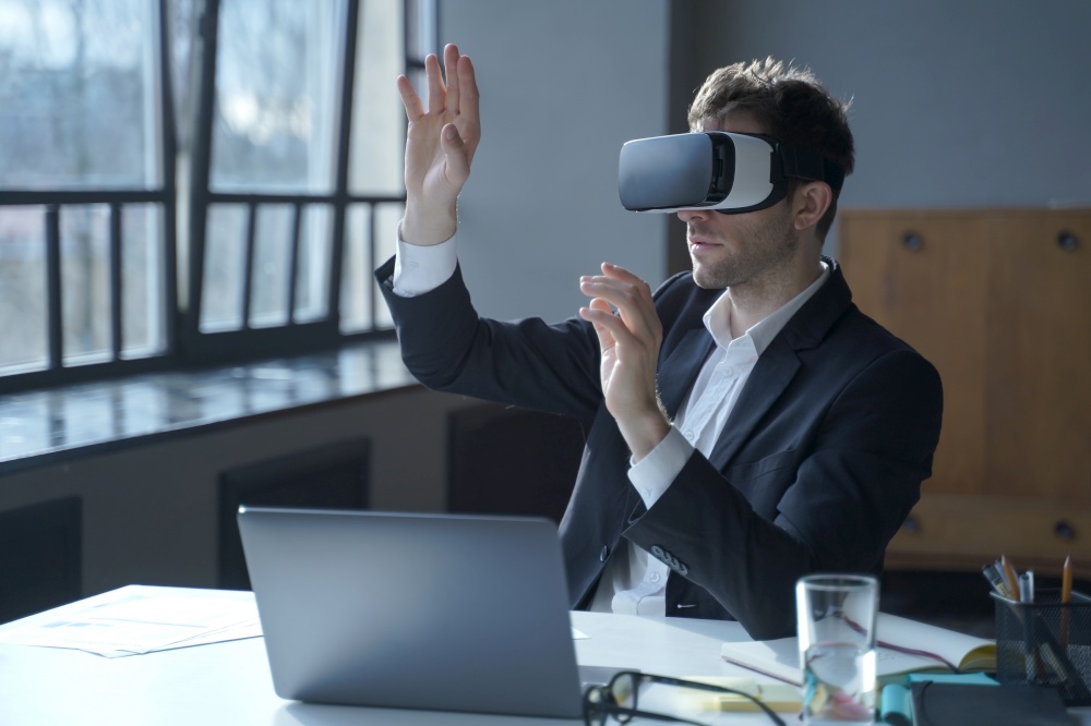 Male office worker in vr headset interacting with digital interface while sitting at desk, touching 3d objects in air, businessman in suit testing virtual reality goggles for business. Male office worker in vr headset interacting with digital interface while sitting at desk